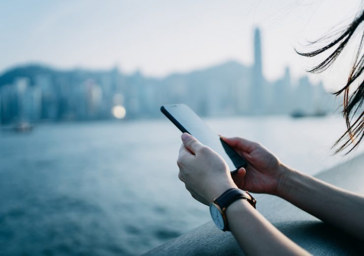 Close up of young woman using mobile phone by the promenade of Victoria Harbour with urban city skyline in background