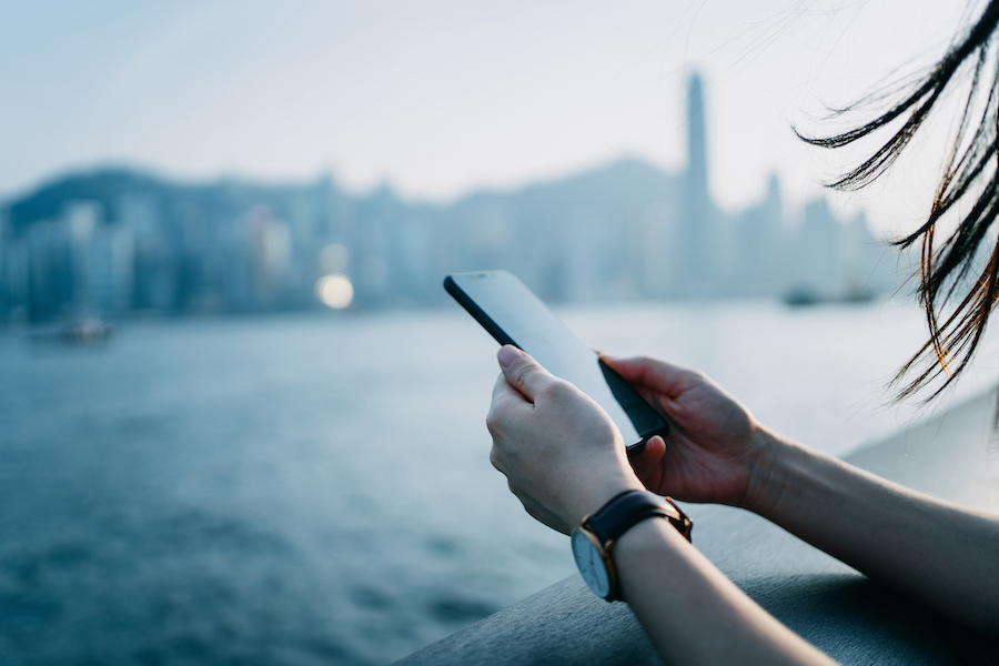 Close up of young woman using mobile phone by the promenade of Victoria Harbour with urban city skyline in background